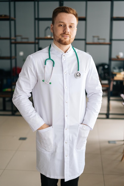 Vertical portrait of smiling male doctor wearing white medical uniform standing with stethoscope in hospital office looking at camera