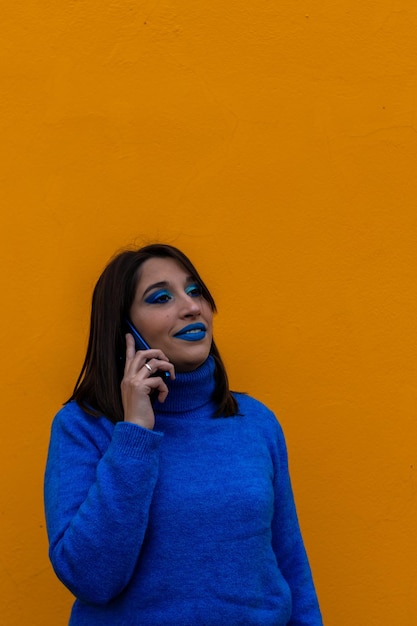 Vertical portrait of smiling young brunette woman in blue dress and makeup talking on the phone on yellow background