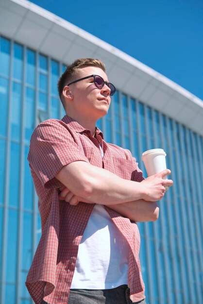 Vertical portrait of stylish handsome young man is drinking coffee or tea from paper cup outdoors at summer sunny day in sunglasses