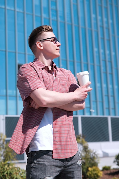 Vertical portrait of stylish handsome young man is drinking coffee or tea from paper cup outdoors at