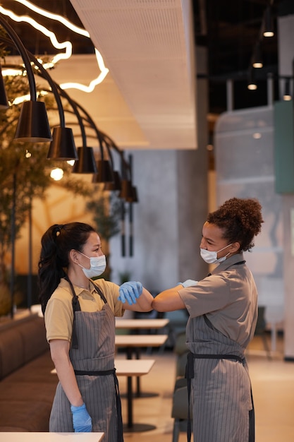 Vertical portrait of two young women bumping elbows as contactless greeting while working in cafe, covid safety concept