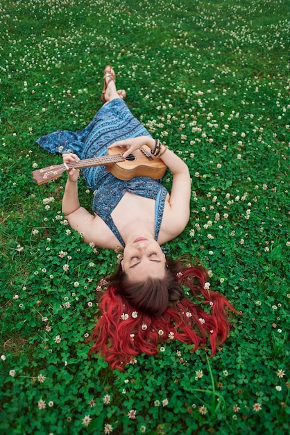 Vertical portrait woman relaxes lying on the grass in summer playing ukulele with closed eyes.