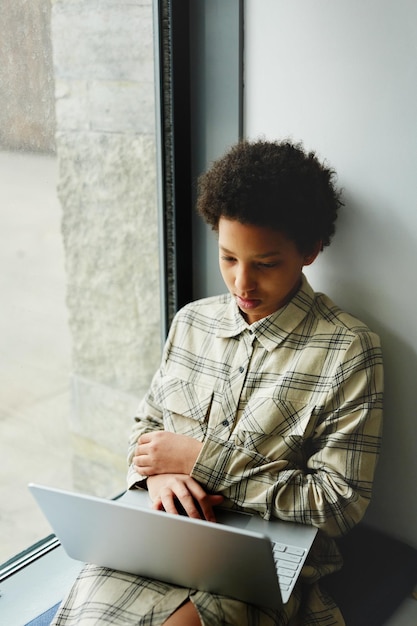 Vertical portrait of young black girl using laptop and watching videos while sitting by window in sc