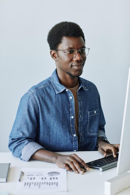 Vertical portrait of young black professional at workplace in office using pc computer against white