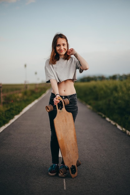 Vertical poster portrait young woman with tattoo leaning on a longboard looks into the camera woman standing in the middle of the road