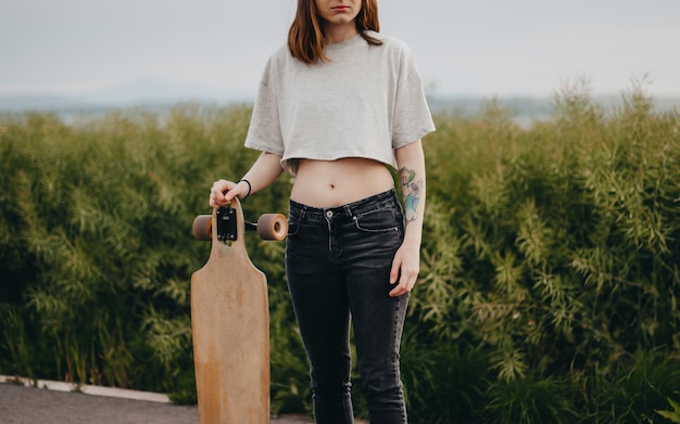 Vertical poster young faceless woman holding a longboard in her hand in the background is green natu