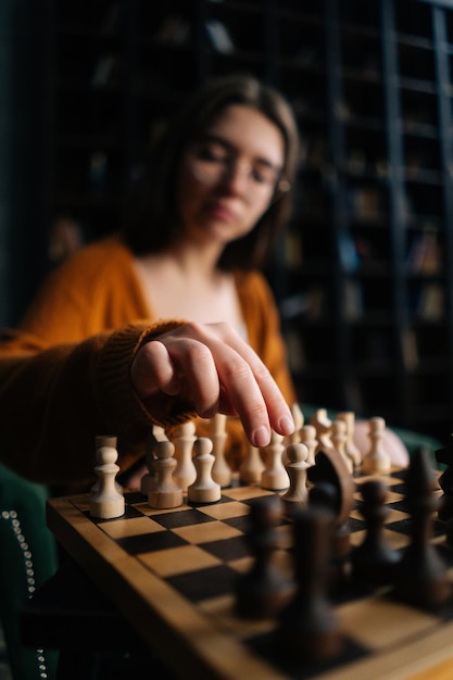 Vertical selective focus shot of young woman wearing elegant eyeglasses making chess move sitting on armchair in dark room