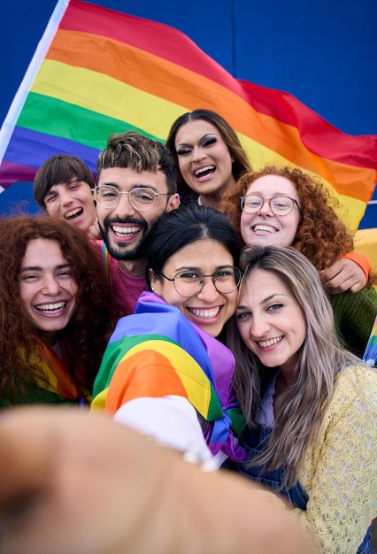 Vertical selfie of lgbt group young people celebrating gay pride day holding rainbow flag together