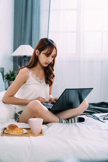 vertical shot attractive asian lady working from home on the laptop with concentration while having breakfast on bed in a white bedroom.