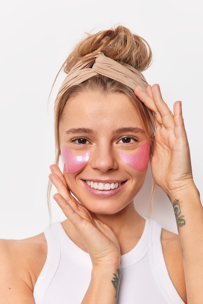 Vertical shot of beautiful cheerful young woman applies hydrogel patches under eyes to reduce wrinkles smiles toothily wears headband isolated over white background. Skin care and beauty concept