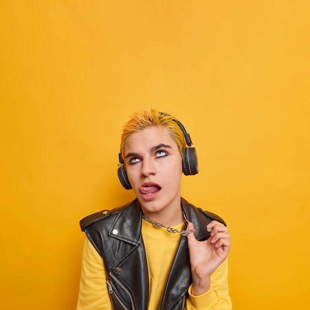 Vertical shot of bored punk girl sticks out tongue touches metal chain concentrated above listens music in wireless headphones wears leather jacket poses against yellow with blank space