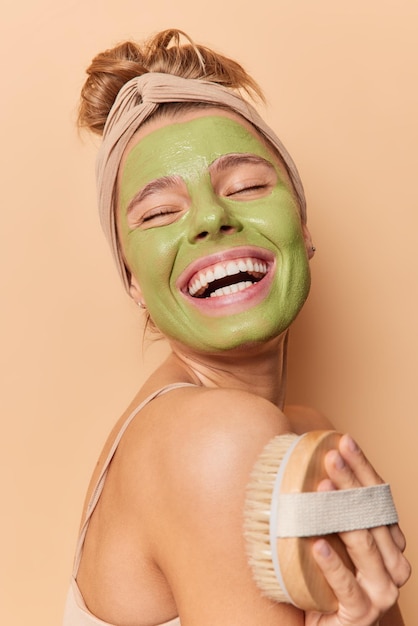 Vertical shot of cheerful young woman laughs happily uses body brush applies green facial mask wears headband poses against brown background feels very happy. Skin care and treatment concept