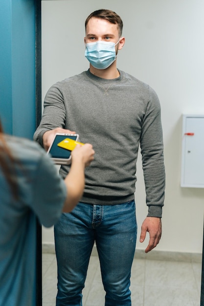 Vertical shot of courier male in protective face mask giving POS wireless terminal to making contactless payment via credit card