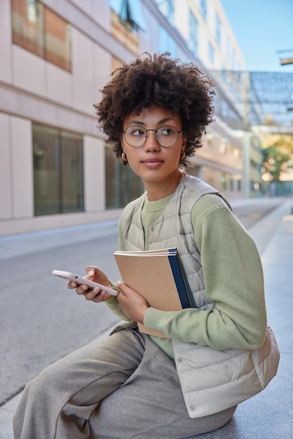 Vertical shot of curly haired young woman waits for someone outdoors uses mobile phone for chatting online holds textbooks