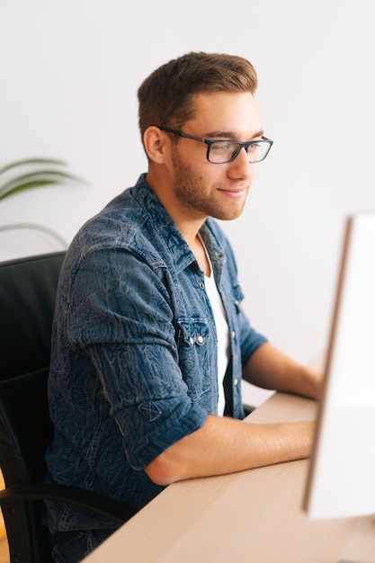 Vertical shot of handsome young designer male in glasses working on desktop computer sitting at desk at home office in room with modern interior