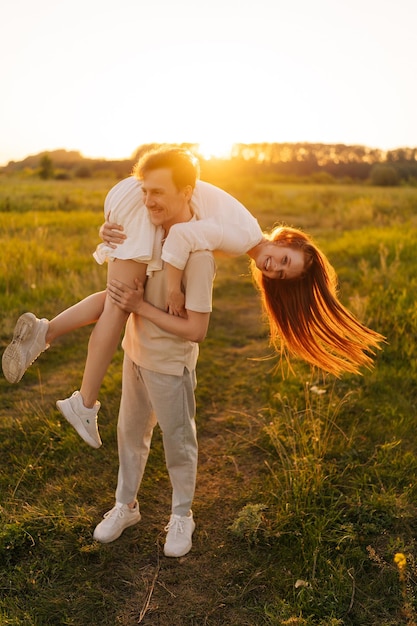 Vertical shot of happy young married couple in love playing enjoying on green field during golden sunset on summer evening