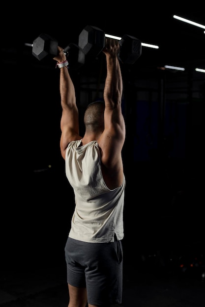 Vertical shot of a muscular man from the side pressing a pair of dumbbells