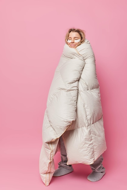 Vertical shot of pleased young woman wrapped in comfy blanket keeps eyes closed applies beauty patches under eyes for skin treatment enjoys good rest isolated over pink background Sleeping concept