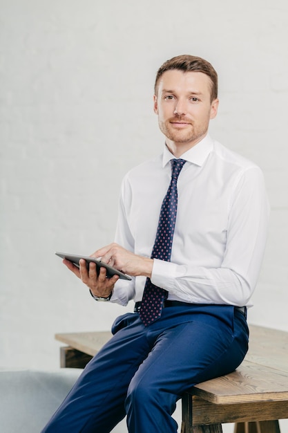 Vertical shot of serious businessman uses modern tablet computer for reading financial news online makes banking browses web page connected to wireless internet People and technology concept