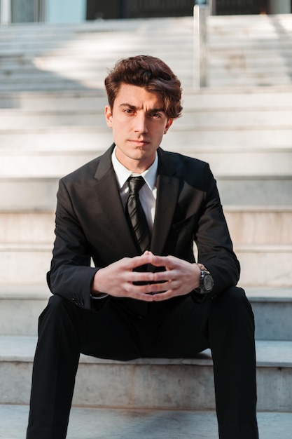 Vertical shot of a serious young Caucasian guy dressed professionally sitting on outdoor stairs