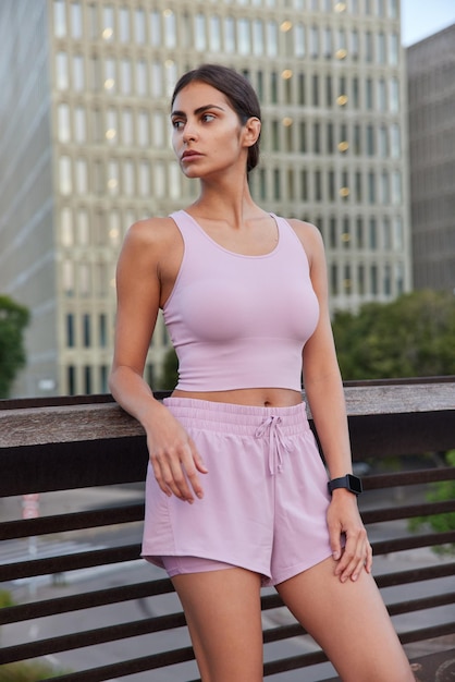 Vertical shot of sporty young woman dressed in cropped top and shorts rests after doing sport exercises looks away poses on bridge against urban background. Active female model has morning training