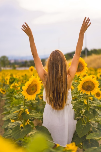 Vertical shot of a young blonde with a white dress walking in the sunflower field