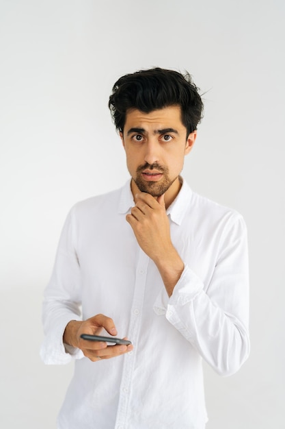 Vertical studio portrait of thinking young man in casual shirt looking at camera holding smartphone standing on white isolated background