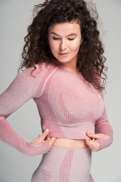 Vertical view of adult woman with fit body wearing pink sports bra and shorts standing alone at the studio. Female straightens her clothes. Sport concept