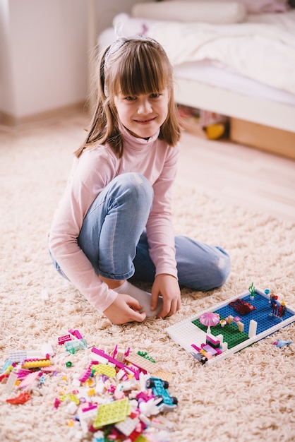 Photo vertical view of pretty smiling little toddler girl sitting on the carpet of her room and looking at the camera while playing with plastic toys.