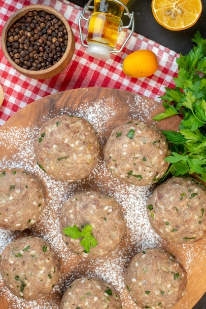 Vertical view of uncooked meat balls with green in a brown plate and rice fresh green lemon fallen oil bottle pepper on red stripped towel on black background