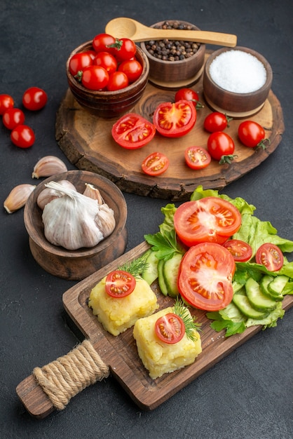 Vertical view of whole cut fresh vegetables and spices on wooden board white towel cutlery set cheese on black surface