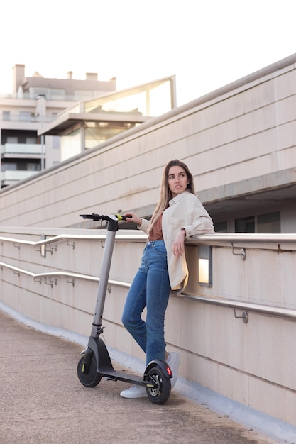 Vertical view of young woman leaning resting with her electric scooter wating for her friend to come with sun behind