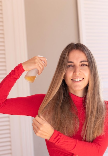 Vertical view of young woman looking to camera applying spray treatment in the shiny and beautiful blond long hair