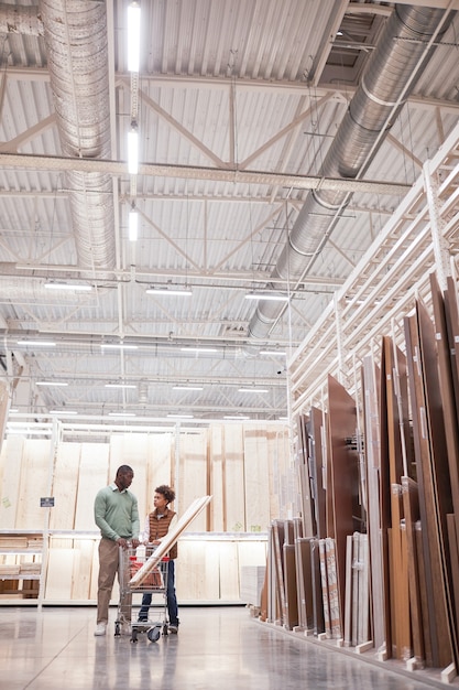 Vertical wide angle portrait of African-American father and son shopping together in hardware store standing with cart in wood and boards isle, copy space