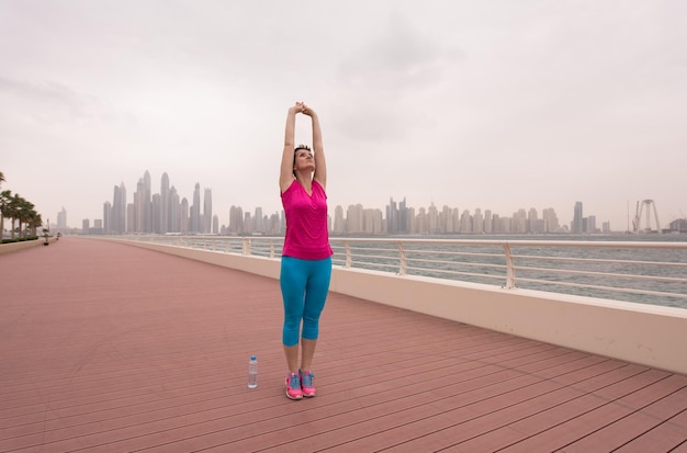 very active young beautiful woman stretching and warming up on the promenade along the ocean side with a big modern city in the background to keep up her fitness levels as much as possible