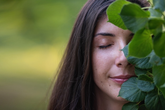 Very closeup portrait of a freckled young woman covering part of her face with green linden leaves