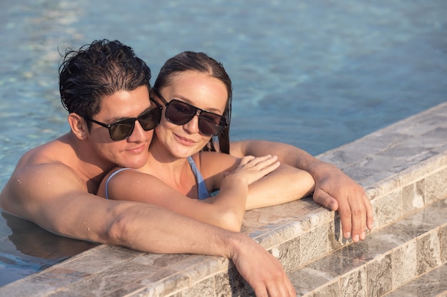 Very happy couple wearing sunglasses enjoy each other in swimming pool. Man holding woman on hands