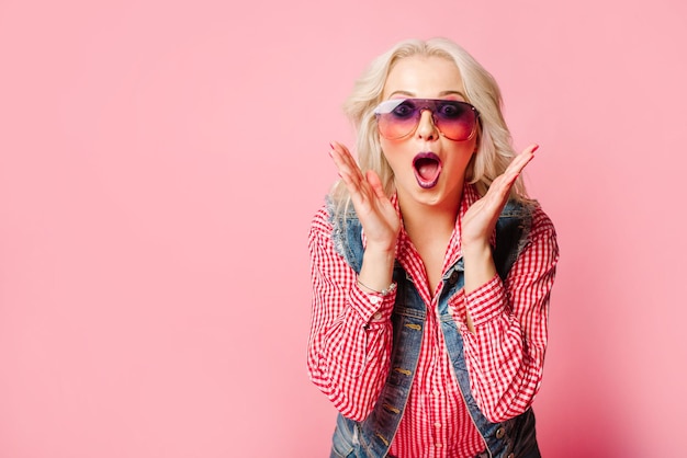 Very positive and happy adult blond woman dressed in shirt and blue jeans posing on pink background