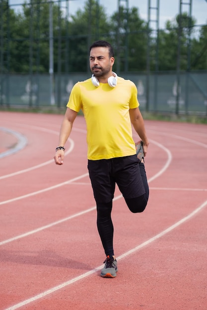 Vibrant athlete in wireless headphones warms up leg muscles on rubberized running track at city park