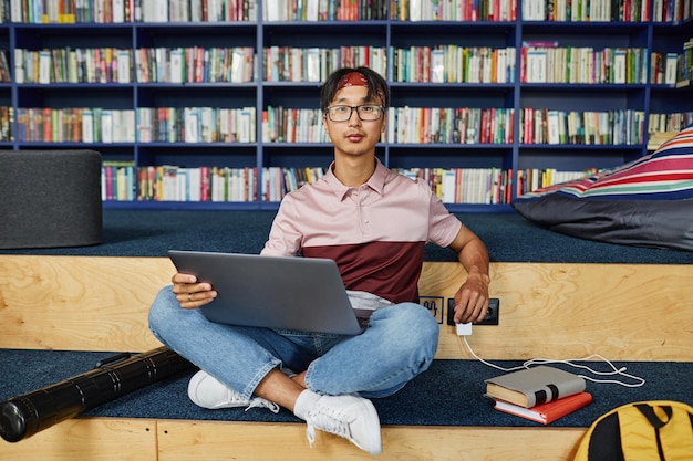 Vibrant full length portrait of young asian man sitting cross legged with laptop in college library