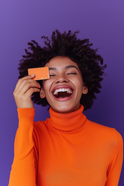 Vibrant and joyful woman in an orange turtleneck laughing and holding a credit card against a purp