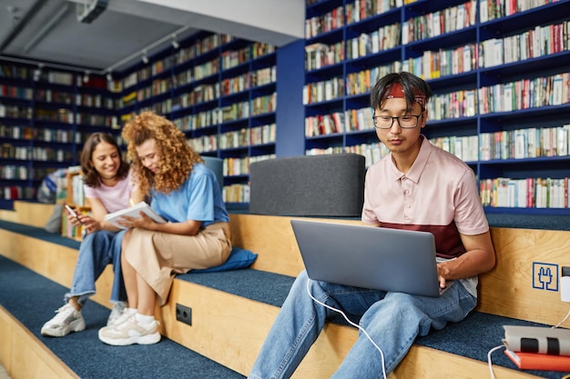 Vibrant portrait of asian young man using laptop in college library and studying with people in back