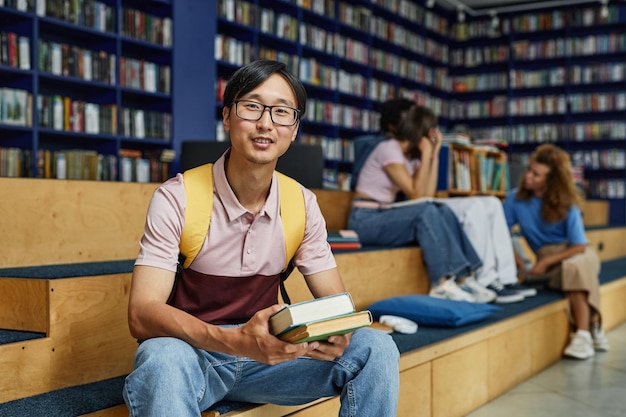 Vibrant portrait of young asian man holding books in college library and smiling at camera copy spac