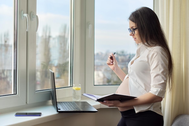 Video communication call pregnant woman with laptop working at home near the window