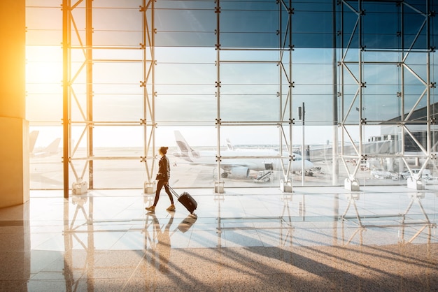 View on the aiport window with woman walking with suitcase at the departure hall during the sunset. Wide angle view with copy space