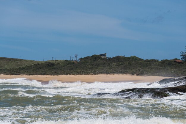 View of Areia Negras beach in Rio das Ostras in Rio de Janeiro with sunny day, blue sky and some clouds. Strong sea and black sand mixed with yellow and many rocks.