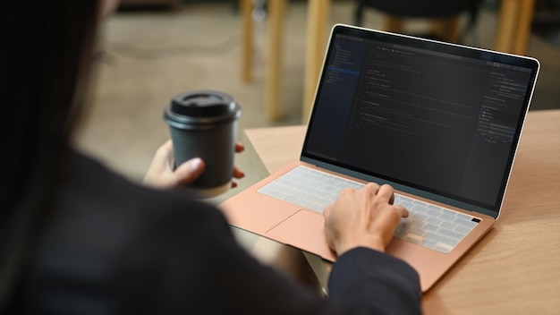 View over businesswoman shoulder holding paper cup and using laptop computer at her workplace