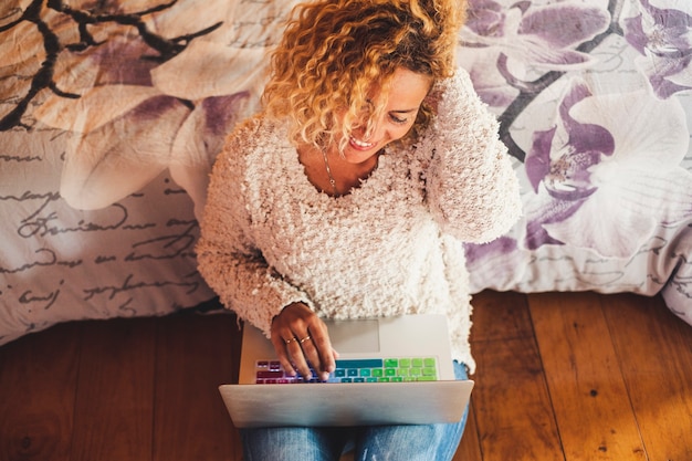 Above view cheerful caucasian curly woman working on laptop computer sitting on the wooden floor