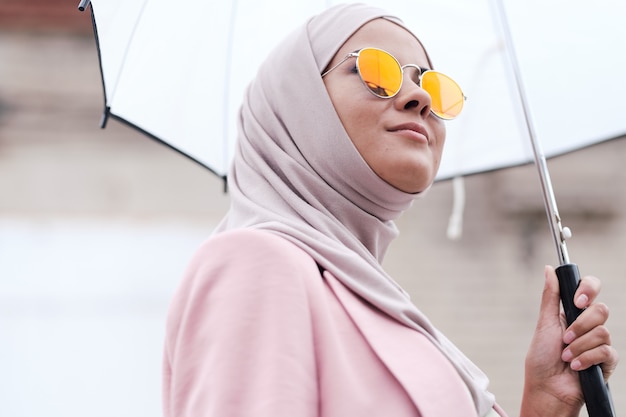 Below view of content confident young Muslim woman in sunglasses and hijab enjoying stroll under rain