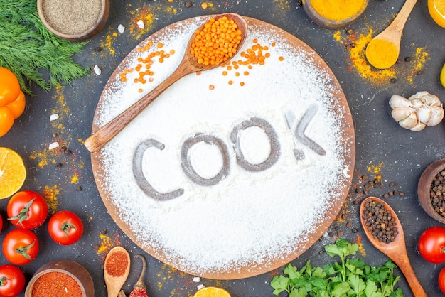 Above view of cook writing with flour on wooden board among fresh vegetables spices green bundles on black background